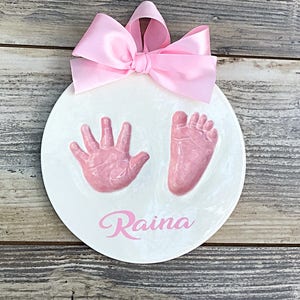 Op de afbeelding: Ronde witte keramische hanger met roze hand- en voetafdrukken en de naam "Raina" in roze cursief.