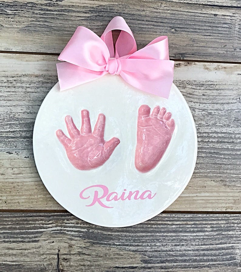 Op de afbeelding: Ronde witte keramische hanger met roze hand- en voetafdrukken en de naam "Raina" in roze cursief.