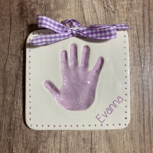May include: A white ceramic handprint plaque with a purple handprint and the name "Evanna" written in purple lettering. The plaque has a purple and white gingham ribbon tied around the top.