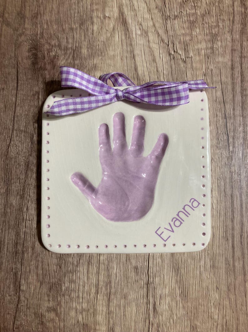 May include: A white ceramic handprint plaque with a purple handprint and the name "Evanna" written in purple lettering. The plaque has a purple and white gingham ribbon tied around the top.