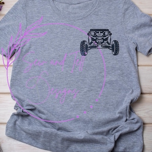 Puede incluir: Camiseta gris con un gráfico negro de un vehículo todoterreno. La camiseta tiene cuello redondo y mangas cortas. El texto "Saw and 14 Designs" está impreso en rosa en una fuente cursiva dentro de un círculo rosa.