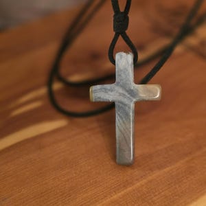 Może przedstawiać: Wisiorek w kształcie krzyża wykonany z gładkiego, polerowanego kamienia z wirującymi szarymi, białymi i brązowymi wzorami. Krzyż zawieszony jest na prostym czarnym sznurku przechodzącym przez otwór u góry, tworząc naszyjnik o naturalnym wyglądzie.