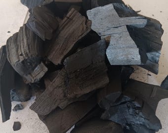 May include: A pile of irregular, black charcoal lumps with visible wood grain textures. A silver-colored United States quarter coin is placed next to the charcoal for scale, measuring approximately 0.955 inches in diameter.