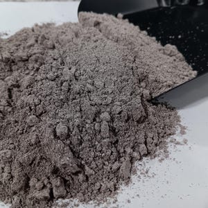 May include: A pile of fine, dark gray powder with a slightly reddish hue is shown. A black scoop is partially visible, suggesting the powder is being measured or dispensed. The powder appears to be a dry, granular substance.