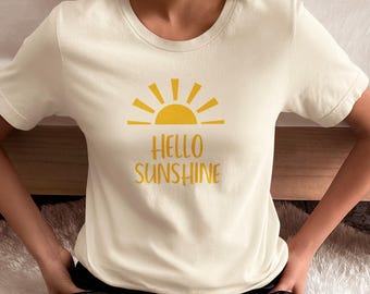May include: Cream-colored t-shirt with the text "HELLO SUNSHINE" in yellow, featuring a yellow sun graphic. The shirt is short-sleeved and has a round neckline. The word "Natural" is in a black circle in the upper left corner.