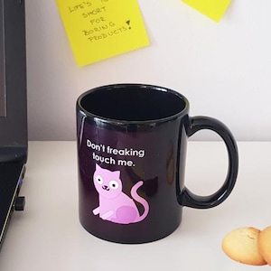 Op de afbeelding: Zwarte koffiemok met een roze kattenillustratie en de tekst "Don't freaking touch me."