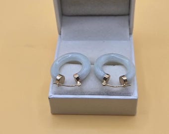 May include: A pair of light green jade hoop earrings with gold-colored clasps, presented in a gray jewelry box. The earrings have a smooth, rounded design and a subtle, translucent quality. The box is open, showcasing the earrings against a neutral background.