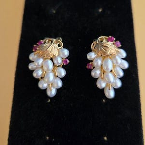 May include: A pair of gold-toned earrings shaped like grape clusters. Each earring features a cluster of white pearls, accented with small red gemstones and a gold leaf design at the top. The earrings are displayed on a black surface.