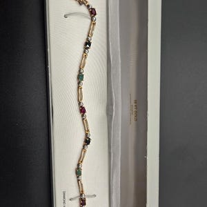 May include: A gold-toned bracelet with alternating gemstones of red, green, and clear stones, presented in a white jewelry box. The bracelet is rectangular in shape and has a clasp closure. The box is open, revealing the bracelet against a white satin lining.