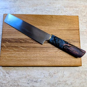 Può includere: Un coltello da chef artigianale con una lama in acciaio lucido è appoggiato su un tagliere rettangolare in legno. Il coltello presenta un manico con tonalità blu e viola. Il tagliere è in legno marrone chiaro.
