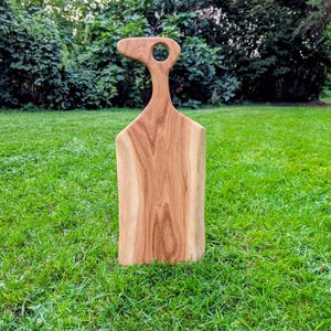 Könnte beinhalten: Ein Holzschneidebrett mit Griff und kreisförmiger Öffnung. Das Brett besteht aus hellem Holz und hat eine natürliche, organische Form. Das Brett steht auf grünem Gras.