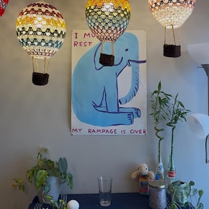May include: Three crocheted hot air balloons hanging from the ceiling. The balloons are in shades of yellow, red, and white. The balloons are hanging over a blue poster with a cartoon elephant and the text "I must rest my rampage is over".