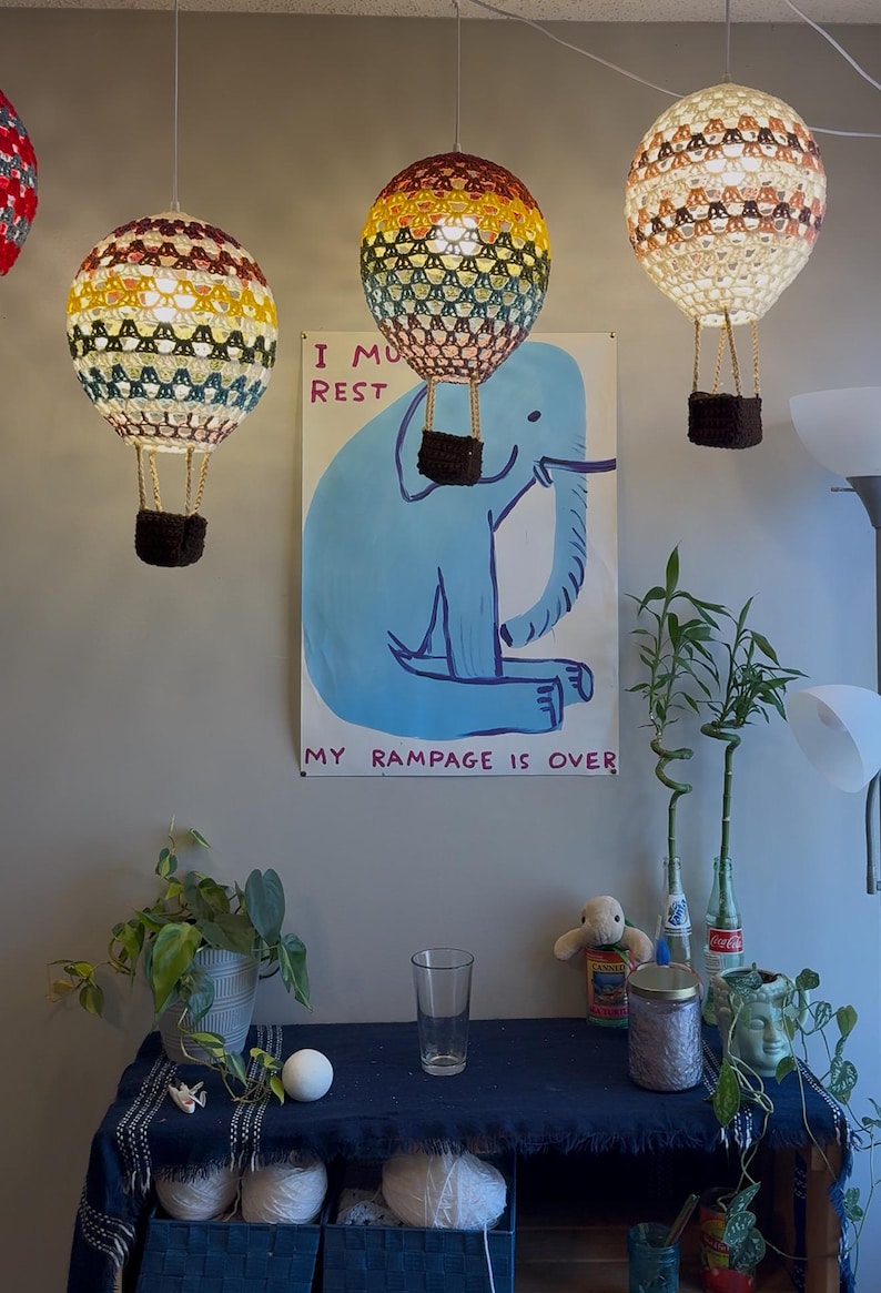 May include: Three crocheted hot air balloons hanging from the ceiling. The balloons are in shades of yellow, red, and white. The balloons are hanging over a blue poster with a cartoon elephant and the text "I must rest my rampage is over".
