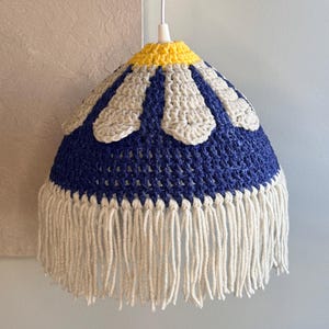 May include: A handmade crochet lampshade in a daisy design. The lampshade features a navy blue body with white and yellow accents. The bottom edge is trimmed with long, cream-colored fringe. The lampshade hangs from a white cord.