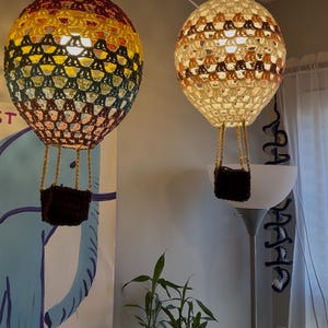 May include: Two crocheted hot air balloon light fixtures hanging from the ceiling. The balloon on the left is multi-colored with yellow, orange, red, green, and brown. The balloon on the right is cream colored with brown and orange accents.