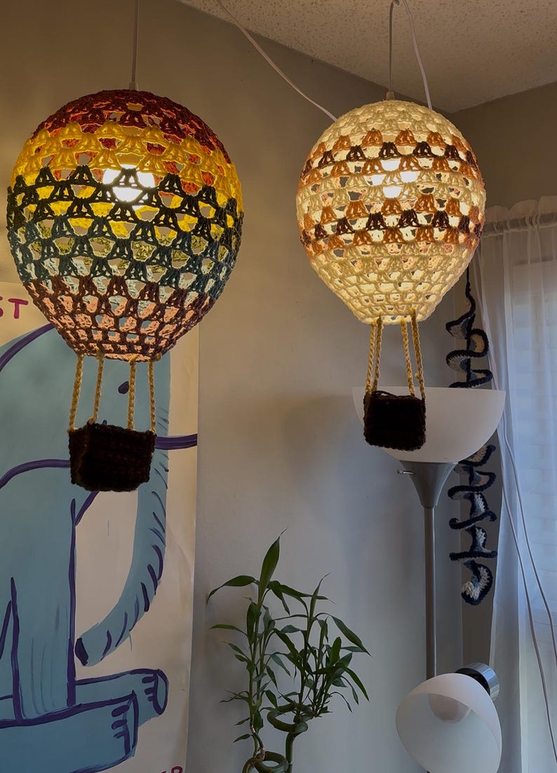 May include: Two crocheted hot air balloon light fixtures hanging from the ceiling. The balloon on the left is multi-colored with yellow, orange, red, green, and brown. The balloon on the right is cream colored with brown and orange accents.