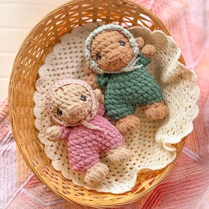 May include: Two handmade crochet dolls in a woven basket. One doll is wearing a pink outfit and the other is wearing a green outfit. Both dolls have brown skin and are wearing bonnets. The basket sits on a cream-colored crocheted doily.