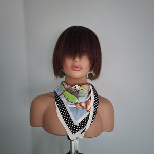 Peut inclure: Une tête de mannequin avec une perruque courte brun foncé, portant un foulard blanc et noir à pois avec un imprimé coloré. Le foulard est noué autour du cou et les extrémités pendent. Le mannequin est sur un trépied.