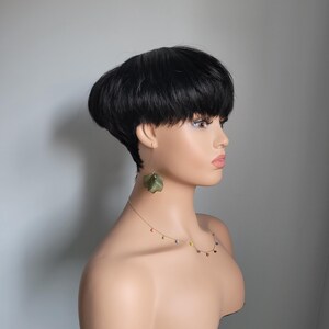 May include: A black wig with a short, rounded, bowl-shaped cut. The wig is styled with a side parting and is worn on a mannequin head. The mannequin is wearing a gold necklace with colourful beads.