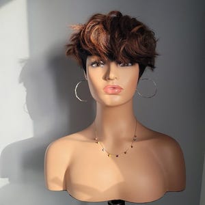 May include: A mannequin head with short, brown and blonde wavy hair, wearing silver hoop earrings and a delicate necklace with colourful beads. The mannequin is mounted on a black tripod.