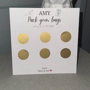Op de afbeelding: Een witte kaart met de tekst "AMY Pack your bags MATCH 3 TO WIN" en zes gouden krascirkels. De kaart bevat ook een papieren vliegtuigje en een paspoortafbeelding, met de woorden "Love Mam & Dad" onderaan.