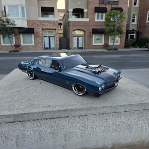 May include: A dark blue classic car model with chrome accents and a prominent engine. The car is parked on a concrete surface, showcasing its sleek design and detailed features. The model is a vintage muscle car.