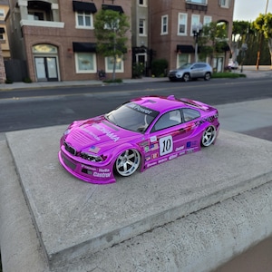 Może przedstawiać: Żywy, magentowy samochodzik z naklejkami wyścigowymi i numerem 10 z boku. Samochód ma srebrne felgi i jest zaparkowany na betonowej powierzchni. Na samochodzie widnieją napisy "Yokohama", "Hella" i "Castrol".