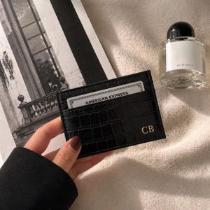 May include: Black cardholder with a crocodile texture, holding an American Express card. The cardholder has gold initials "CB" and is next to a perfume bottle.