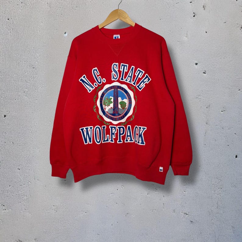 Op de afbeelding: Een rode sweatshirt met de tekst "N.C. STATE WOLFPACK" in witte en blauwe letters. De sweatshirt heeft een cirkelvormig embleem met een vredesteken en een landschapsontwerp. De sweatshirt hangt aan een houten hanger.