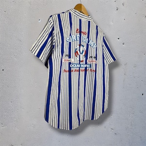 May include: A short-sleeved, button-down shirt with vertical blue and white stripes. The back of the shirt features the text "Surf Camp" in blue, along with a graphic of a life preserver and the words "Ocean Pacific".