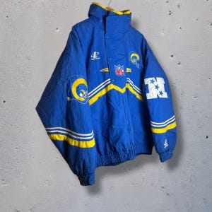 May include: Blue and yellow vintage jacket with the Los Angeles Rams logo and NFL branding. The jacket features a hood, white stripes, and the number "4" on the sleeve. The jacket is made of a lightweight material.