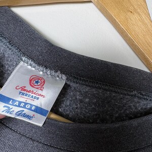 Op de afbeelding: Close-up van een donkergrijze sweatshirt met een label met de tekst "American Threads Collection". Het label geeft ook de maat "LARGE" en het merk "The Game" aan.