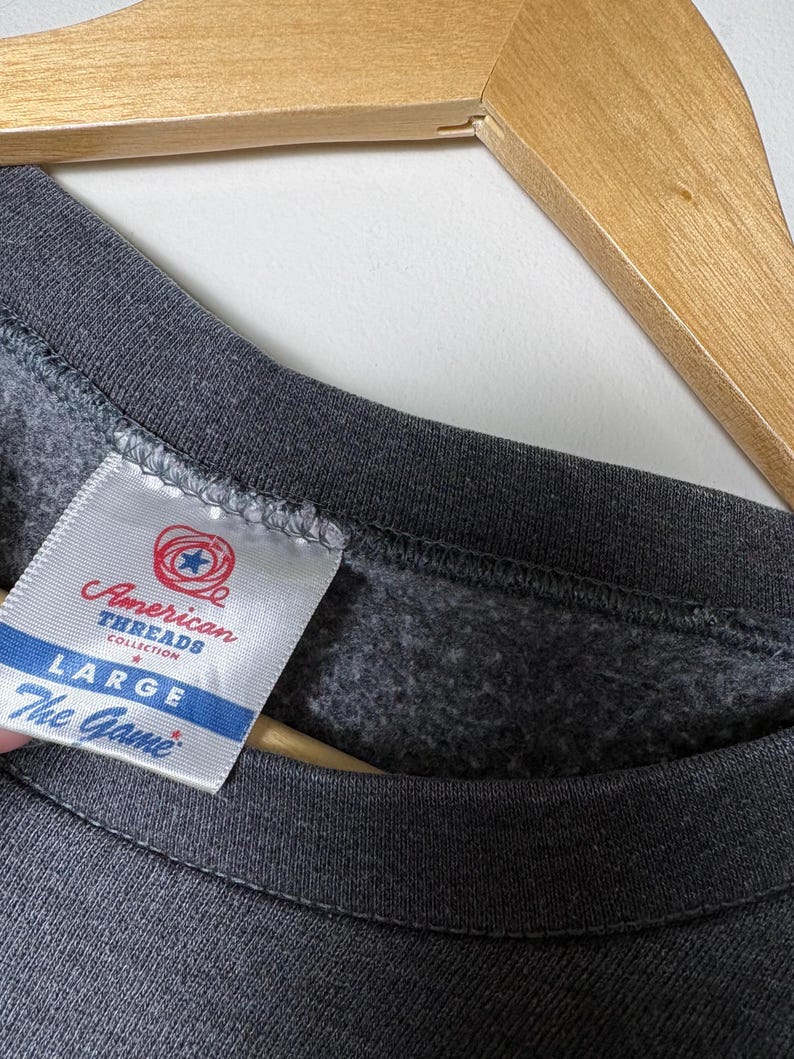 Op de afbeelding: Close-up van een donkergrijze sweatshirt met een label met de tekst "American Threads Collection". Het label geeft ook de maat "LARGE" en het merk "The Game" aan.
