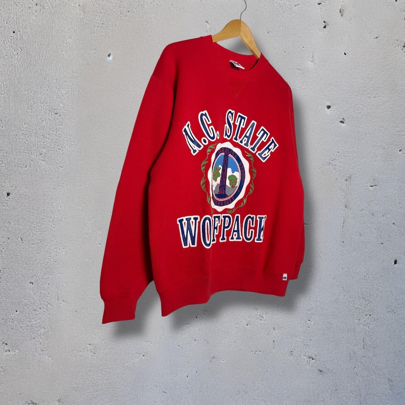 Op de afbeelding: Een rode sweatshirt met de tekst "N.C. STATE" in wit boven een cirkelvormig embleem en het woord "WOLFPACK" in blauw. Het embleem bevat een vredesteken en een boom. De sweatshirt hangt aan een houten hanger.