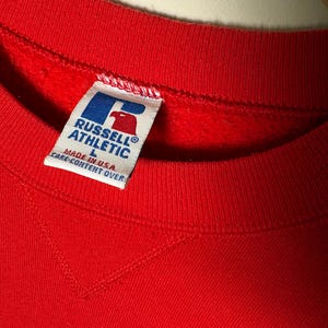 Op de afbeelding: Een close-up van een rode sweatshirt met een ronde hals. Het label luidt "RUSSELL ATHLETIC" met een logo en "MADE IN U.S.A.". De sweatshirt is maat L.
