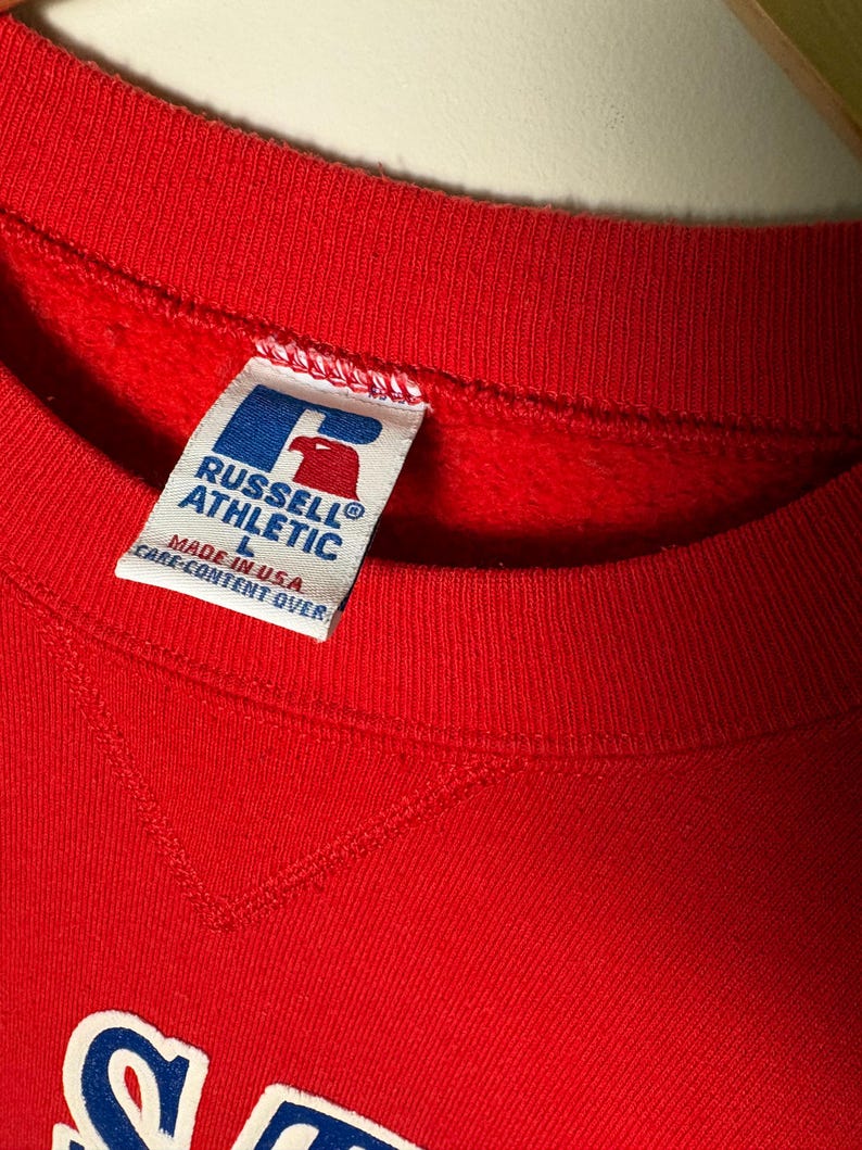 Op de afbeelding: Een close-up van een rode sweatshirt met een ronde hals. Het label luidt "RUSSELL ATHLETIC" met een logo en "MADE IN U.S.A.". De sweatshirt is maat L.