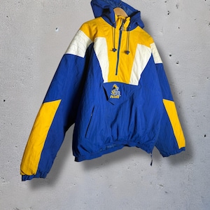 Puede incluir: Chaqueta anorak con capucha azul, amarilla y blanca. La chaqueta tiene cierre de media cremallera, un bolsillo delantero y el logotipo de los Rams. Las mangas y los laterales son azules, con detalles amarillos y blancos en los hombros y la capucha.