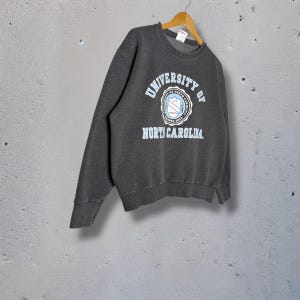 Op de afbeelding: Donkergrijs sweatshirt met de tekst "UNIVERSITY OF NORTH CAROLINA" in lichtblauwe letters. De sweatshirt heeft een ronde hals en lange mouwen. Het embleem van de universiteit staat in het midden.