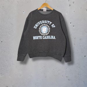 Op de afbeelding: Een donkergrijs sweatshirt met de tekst "UNIVERSITY OF NORTH CAROLINA" in lichtblauwe letters. Het sweatshirt heeft een cirkelvormig embleem met de naam van de universiteit en een schildontwerp. Het kledingstuk hangt aan een houten hanger.