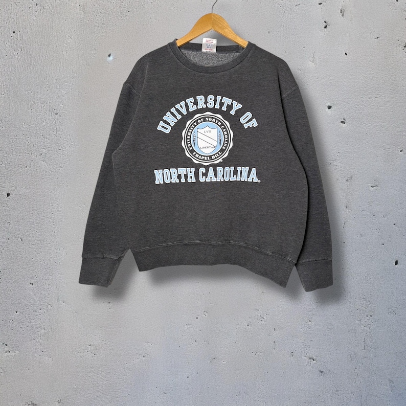 Op de afbeelding: Een donkergrijs sweatshirt met de tekst "UNIVERSITY OF NORTH CAROLINA" in lichtblauwe letters. Het sweatshirt heeft een cirkelvormig embleem met de naam van de universiteit en een schildontwerp. Het kledingstuk hangt aan een houten hanger.