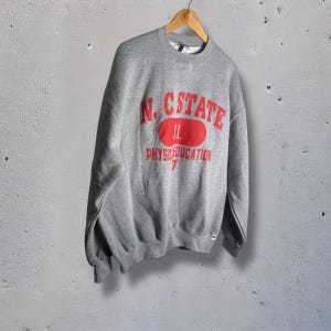 K&ouml;nnte beinhalten: Ein graues Sweatshirt mit dem roten Text "N.C. STATE" &uuml;ber einem roten Oval mit "IL" darin. Unter dem Oval steht "PHYS.EDUCATION" und die Zahl "7". Das Sweatshirt h&auml;ngt an einem Holzb&uuml;gel.