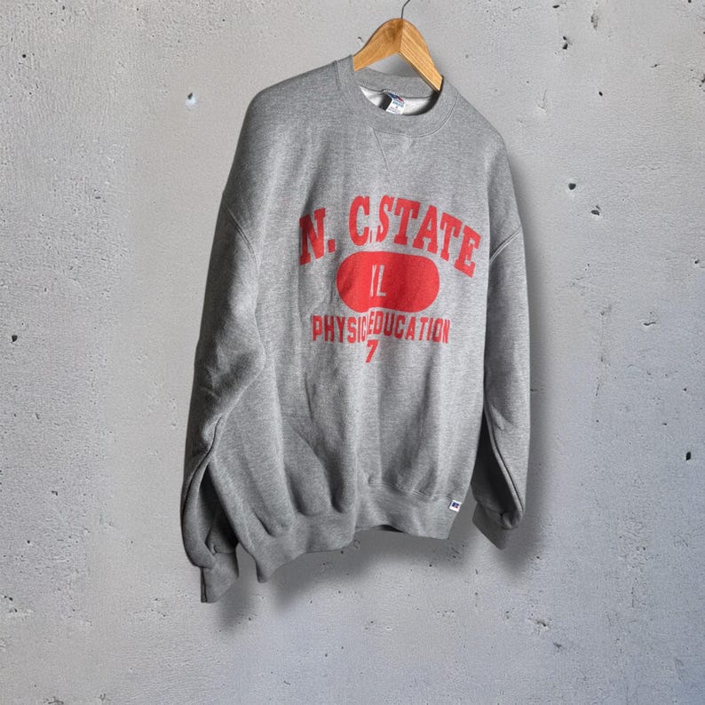 K&ouml;nnte beinhalten: Ein graues Sweatshirt mit dem roten Text "N.C. STATE" &uuml;ber einem roten Oval mit "IL" darin. Unter dem Oval steht "PHYS.EDUCATION" und die Zahl "7". Das Sweatshirt h&auml;ngt an einem Holzb&uuml;gel.