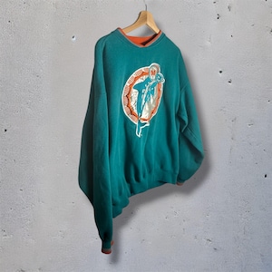 Peut inclure: Sweat-shirt de couleur sarcelle avec un col et des poignets orange. Le devant présente un logo circulaire avec un dauphin bondissant en blanc et sarcelle, souligné d'orange. Le sweat-shirt est suspendu à un cintre en bois.