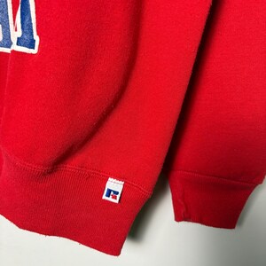 Op de afbeelding: Close-up van een rode sweatshirt met een wit en blauw letterontwerp. De sweatshirt heeft een geribde manchet en een klein wit label met een logo. De stof lijkt een zacht, gebreid materiaal te zijn.