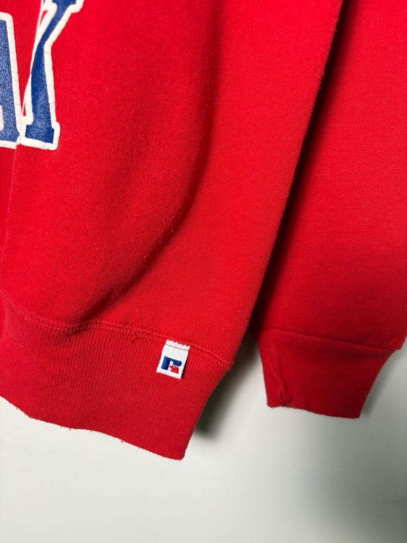 Op de afbeelding: Close-up van een rode sweatshirt met een wit en blauw letterontwerp. De sweatshirt heeft een geribde manchet en een klein wit label met een logo. De stof lijkt een zacht, gebreid materiaal te zijn.