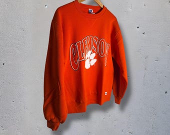 May include: An orange sweatshirt with the word "CLEMSON" in white block letters and a white paw print. The sweatshirt is hanging on a wooden hanger against a gray background.