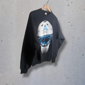 May include: Black sweatshirt featuring a hockey goalie mask graphic. The mask is white with blue and red accents, and the word "RAPS" is visible. The sweatshirt is hanging on a wooden hanger against a gray background.