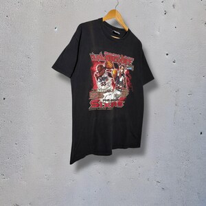 Può includere: T-shirt nero con grafica di basket, la scritta "Dual Autographed Game" e "Sixers". La grafica include immagini di due giocatori di basket. La maglietta è appesa a una gruccia di legno.