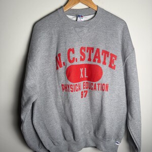 K&ouml;nnte beinhalten: Ein graues Sweatshirt mit Rundhalsausschnitt. Auf der Vorderseite steht in roter Schrift "N.C. STATE" &uuml;ber einem roten Oval mit "XL". Darunter ist "PHYSICAL EDUCATION 97" in Rot gedruckt. Das Sweatshirt h&auml;ngt an einem Holzb&uuml;gel.