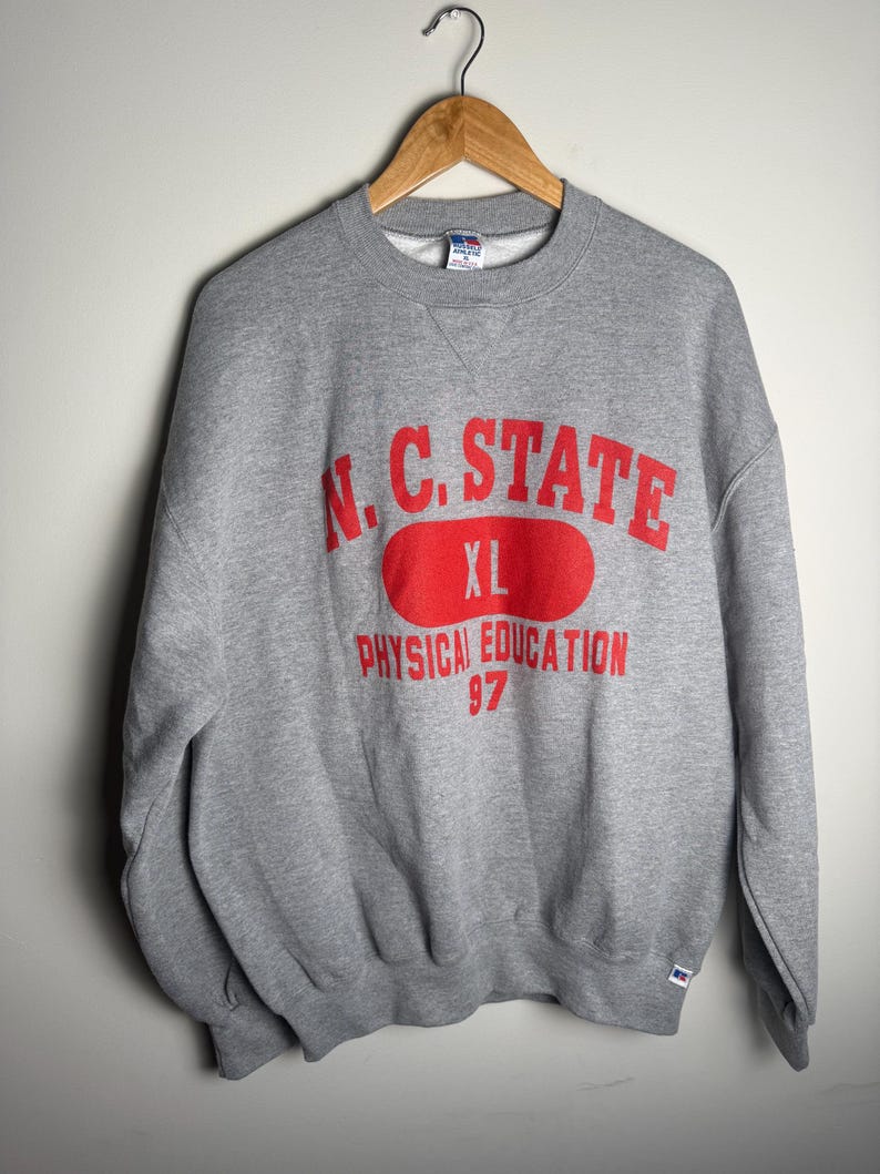 K&ouml;nnte beinhalten: Ein graues Sweatshirt mit Rundhalsausschnitt. Auf der Vorderseite steht in roter Schrift "N.C. STATE" &uuml;ber einem roten Oval mit "XL". Darunter ist "PHYSICAL EDUCATION 97" in Rot gedruckt. Das Sweatshirt h&auml;ngt an einem Holzb&uuml;gel.